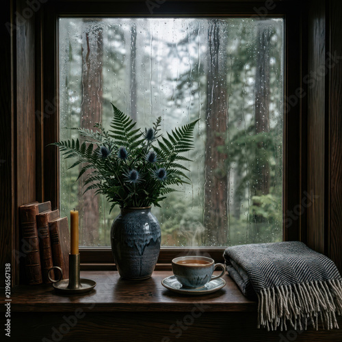 Cozy rainy day view from a wooden cabin window with a steaming cup of tea, books, candle, and a warm blanket