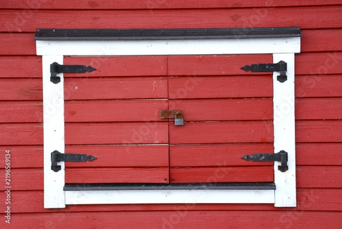 Vue de face d'une fenêtre rectangulaire aux volets rouges en bois fermés, avec un encadrement de couleur blanche. 