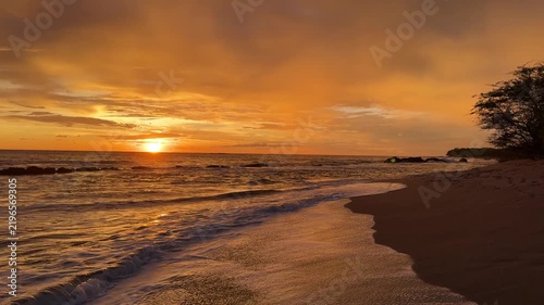 The sun sets over the ocean at a beach location. Waves gently crash on the sandy shore as colors fill the sky.