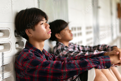 Two depressed teenage boys in plaid shirts sit on the floor of a school hallway, looking sad and lonely.