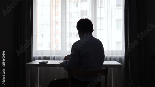 Man in silhouette holding an empty wallet, symbolizing financial crisis, debt, and poverty, sitting at a desk with pieces of paper on it in front of a bright window