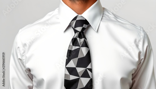 A man in a white shirt and a patterned tie standing against a light background. He has short hair and is looking straight ahead