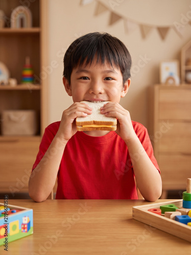 Portrait of a happy Asian boy eating a cheese sandwich and looking directly at camera