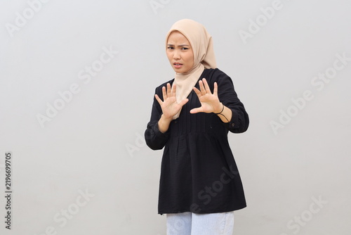Fotografía A young Asian woman wearing a headscarf stands against a white background, displaying an expression of disgust, displeasure, and fear—a reaction of rejection—with her hands raised