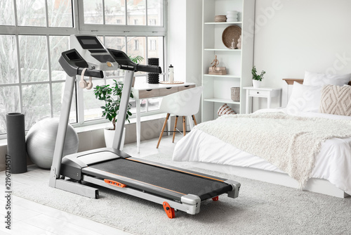 Interior of light bedroom with treadmill, tables and shelf unit