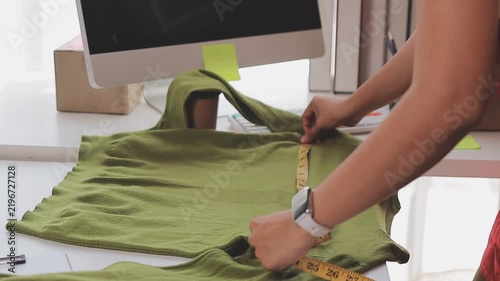 Confident Asian designer woman Beautiful smile using measuring tape for suit jacket with laptop computer in modern studio, clothes hanging on mannequin for customer online sales ordering.