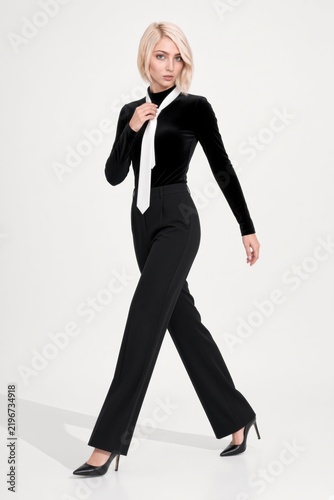 Elegant blonde businesswoman in black attire and white tie walking on a white background.