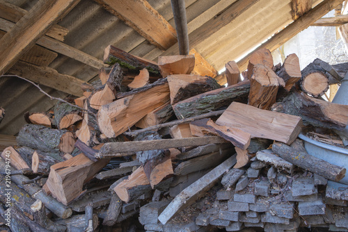 Pile of split firewood and cut logs stored beneath exposed wooden beams in a rustic shed