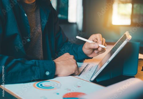 Businessman holding stylus pen and working on tablet screen with financial chart report papers on desk, corporate analytics and digital device concept.