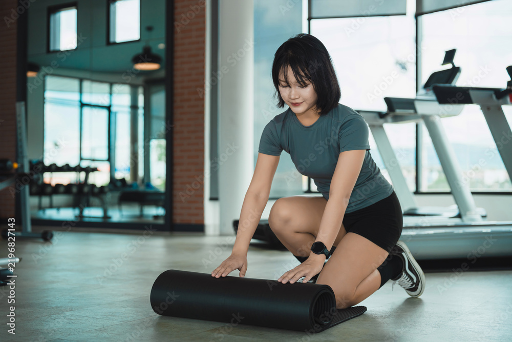 Obraz premium Young woman rolling fitness mat on gym floor preparing for workout session in modern exercise room with equipment