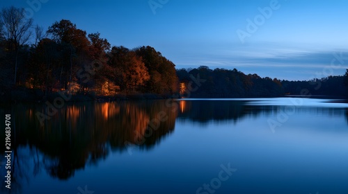 Calm lake with autumn trees illuminated by golden hour light at dusk under blue sky