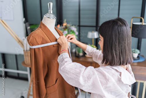Asian woman fashion designer measuring brown suit on mannequin for modern fashion tailoring in creative studio.
