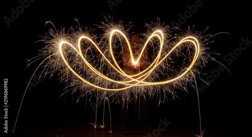 Long exposure light painting of golden sparkler trails forming a symmetrical crown or lotus flower shape held by a person against a black background with glowing sparks.