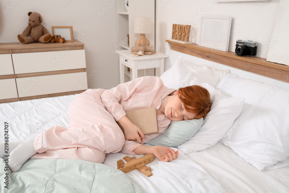 Fototapeta premium Cute teenage happy redhead girl in pajamas with adventure book and wooden airplane sleeping at home