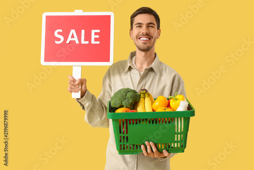 Young man with full shopping basket and SALE sign on yellow background