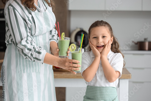 Mother giving glasses of smoothie to surprising little daughter in kitchen