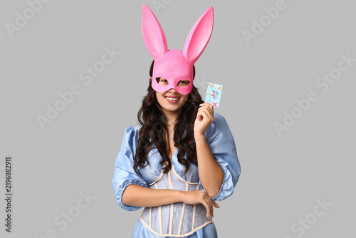 Beautiful young Asian woman in bunny mask with gift card on grey background