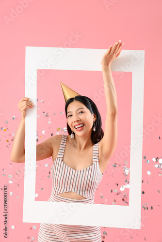 Young beautiful Asian woman in party hat with frame and confetti on pink back...