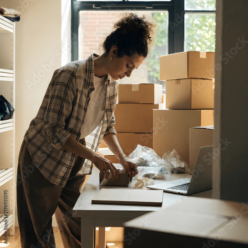 Small business owner packing online store order with cardboard boxes and laptop at workshop desk