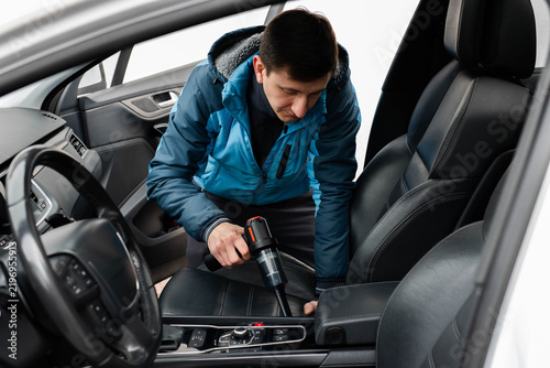 A man is using a compact handheld vacuum cleaner to clean the front leather seats inside a modern car interior