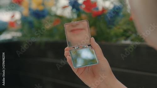 The woman’s mouth is reflected in the pocket mirror she is holding in her hand while sitting on the street.