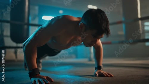 A man performs push-ups in a dimly lit gym with punching bags in the background.
