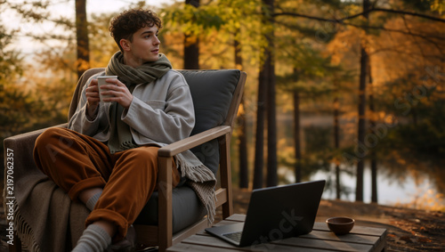 Young man drinking coffee while sitting in an armchair in an autumn forest. Male freelancer with a laptop relaxing outdoors near a lake. Remote work and lifestyle concept