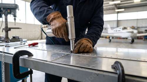 Technician using rivet gun on aircraft metal panel in hangar workshop, with plane visible in background
