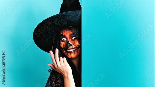 Cheerful witch character leaning around a turquoise backdrop and looking toward the camera, surrounded by bold jack-o-lantern decorations in a playful Halloween scene.