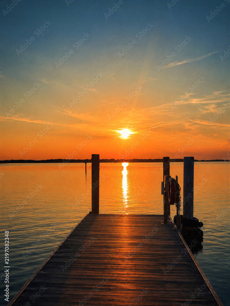 Fototapeta premium Warm sunset reflects across calm water beside a simple wooden pier, creating a peaceful minimalist seascape with glowing sunlight, soft colors, silhouettes, and tranquil evening atmosphere.