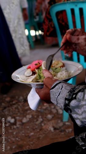 Photography Guest wearing a traditional patterned batik shirt holding a plate of Indonesian festive food with crackers and rice at a social gathering