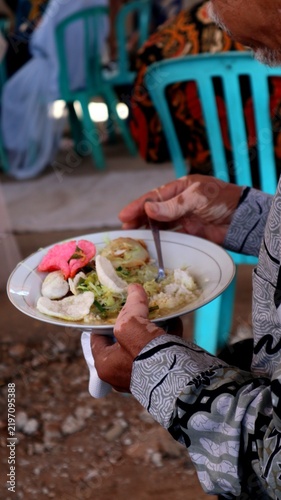 Картината върху платно Guest wearing a traditional patterned batik shirt holding a plate of Indonesian festive food with crackers and rice at a social gathering