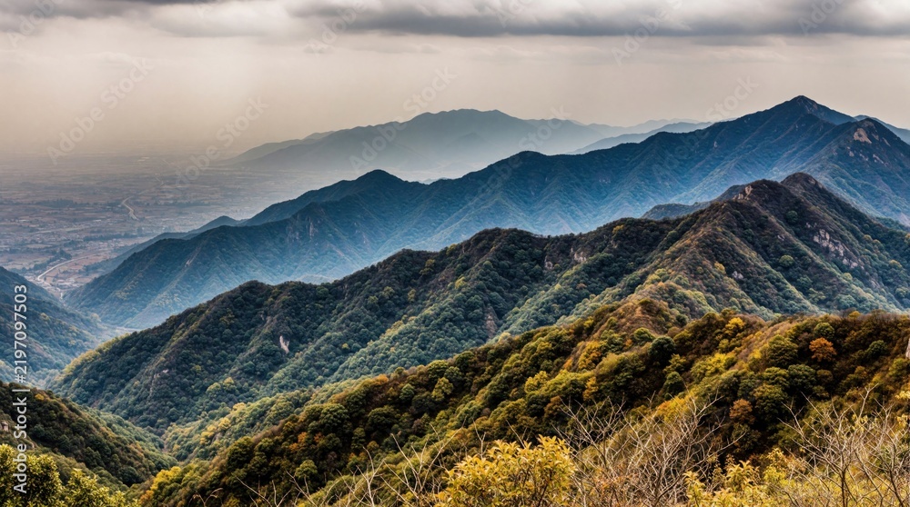 Obraz premium Layers of blue mountain ridges fade into distant haze under cloudy sky. Green forested slopes show autumn colors. Remote valley below is partly visible.