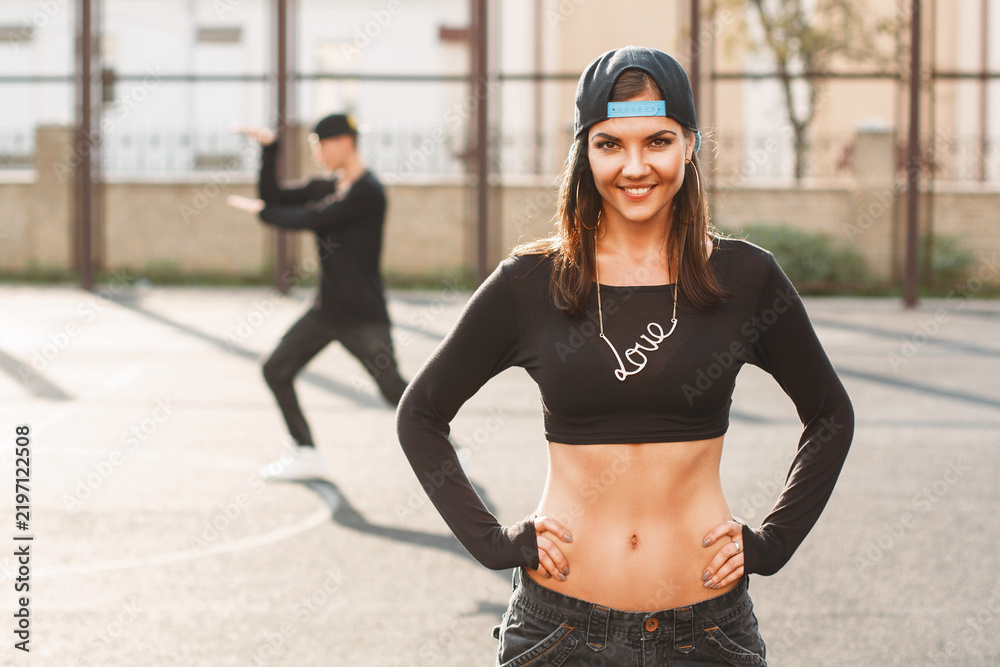 Obraz premium Confident young woman in urban streetwear (crop top, backward cap, 'Love' necklace) poses confidently on an outdoor basketball court. Blurred man dances in background. Bright sun, golden hour light.