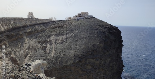 Konstfotografi A dramatic view of a volcanic cliff in Santorini, Greece, showing eroded rock layers and abandoned buildings perched on the edge above the Aegean Sea