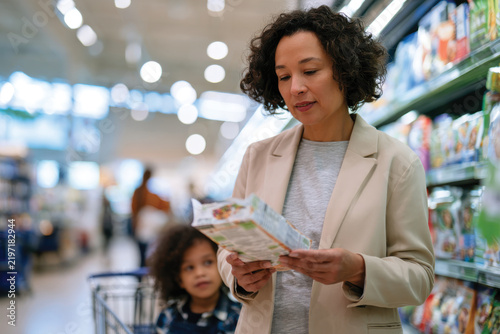 A mother is checking food labels with her child sitting in the shopping cart, representing family life and the importance of healthy food choices in daily routines.