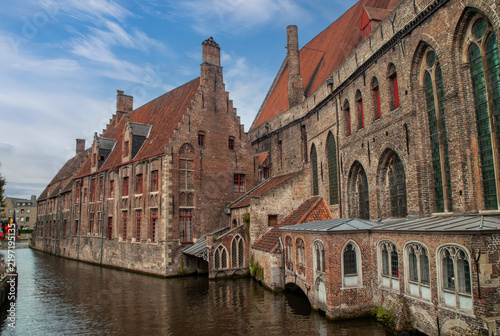 Historic St. John's Hospital beside a canal in Bruges, Belgium, showcasing medieval brick architecture, Gothic windows, red tiled roofs, and the picturesque historic cityscape.