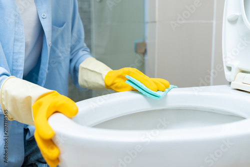 Bild auf Leinwand Close-up of housekeeper wearing yellow rubber gloves cleaning toilet bowl with blue cloth in bathroom