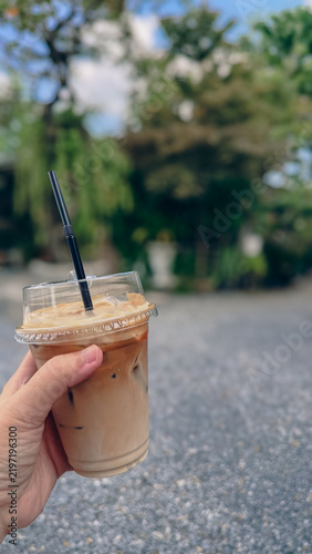Hand holding iced coffee latte in a clear plastic cup outdoors.
