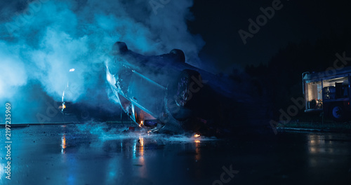 Overturned Car Smolders on the Road After a Severe Traffic Accident at Night, Smoke and Flames Rise From the Wrecked Vehicle as a Fire Truck Stands by at the Car Crash Scene Awaiting Rescue Workers
