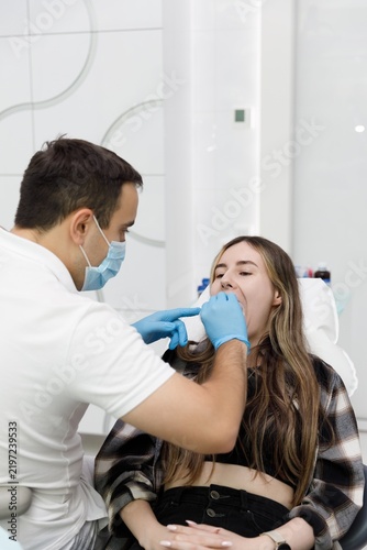 Quadro su tela Dentist placing cheek retractor on smiling female patient in dental chair, dental examination procedure with mouth opener, orthodontic checkup in clinic