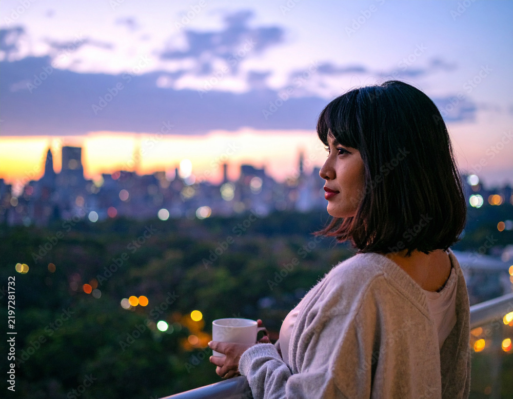 Obraz premium A contemplative woman enjoys a quiet moment at dusk, holding a warm cup while gazing over a blurred city skyline. This serene scene captures urban solitude, peaceful reflection, and evening calm.