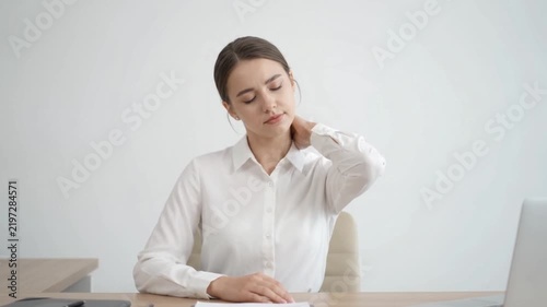 Young woman in a white shirt sitting at a desk and massaging her neck, likely due to pain or stiffness.
