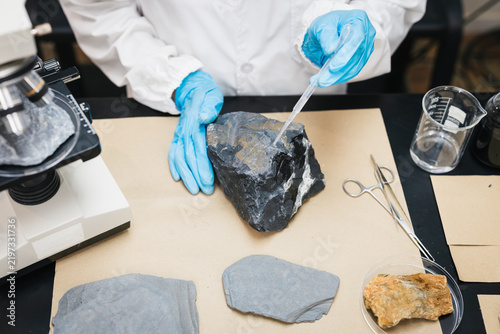 Canvastavla Geologist in a white lab coat and blue gloves examines a dark rock sample with a dropper beside a microscope and mineral specimens