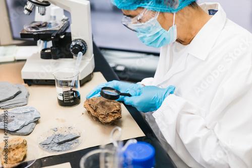 Fotografie Laboratory researcher in protective gear examines a rock sample with a magnifying glass beside a microscope and beakers