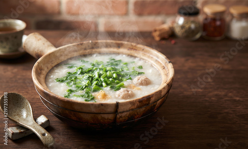 Hot steaming Cantonese congee served in earthenware clay pot with fresh greens pork meatballs wooden table rustic setting comforting warm meal nourishing breakfast culinary tradition