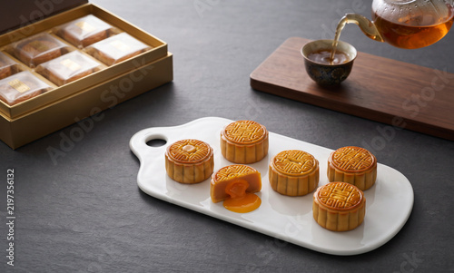 Assorted Chinese mooncakes white ceramic serving tray lava custard filling golden packaging gift box tea pouring ceramic cup dark slate tabletop mid autumn holiday feast   