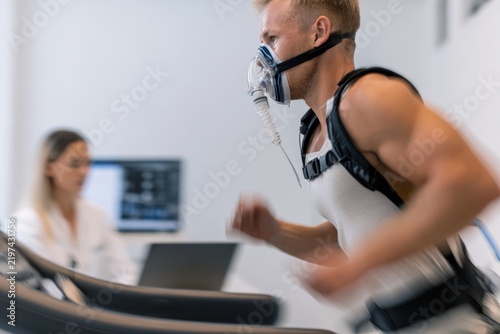 A muscular Caucasian male athlete runs mid-stride on a treadmill while wearing an oxygen mask during a professional physical performance test in a clinic.