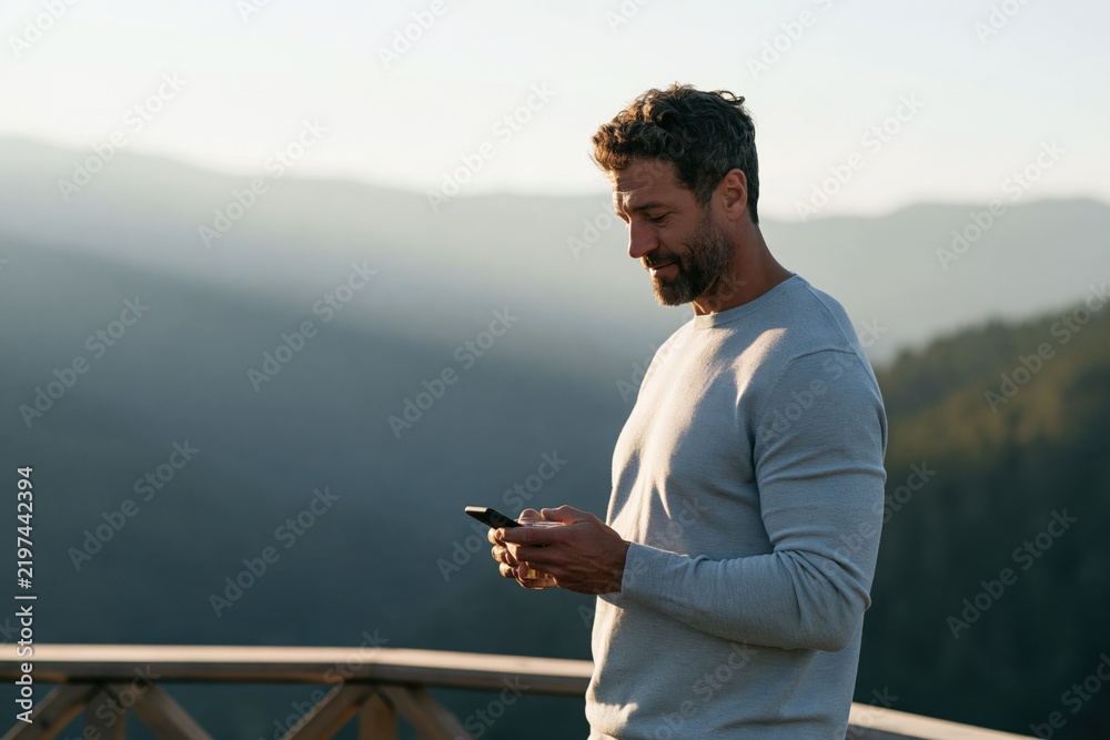Naklejka premium Man checking smartphone on scenic mountain balcony at sunrise for wellness background and website banner