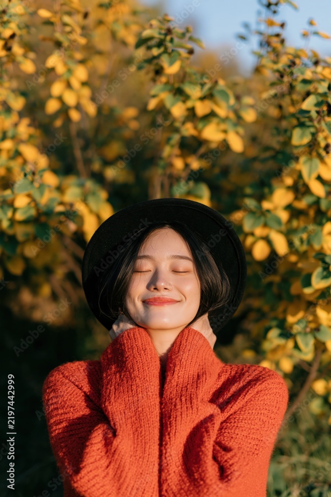 Obraz premium Young woman in a cozy orange sweater and black hat enjoying autumn sunlight among yellow leaves for seasonal portrait background and lifestyle design